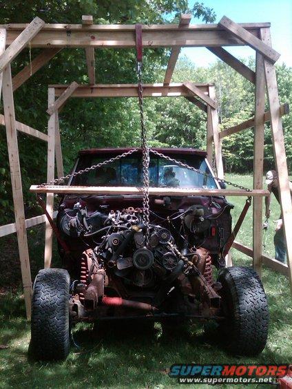 Maroon truck with engine being hoisted out by a wooden frame. Front view, rusty, missing hood