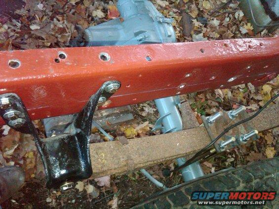 Red painted truck frame rail with black leaf spring bracket and bolts. Gray transmission or transfer case visible behind