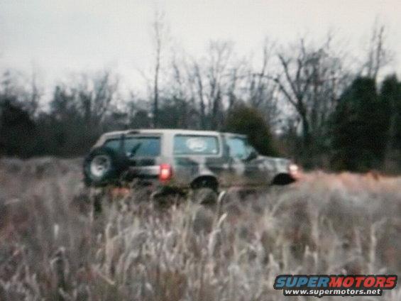1995 Ford Bronco driving through tall dry grass with trees in background