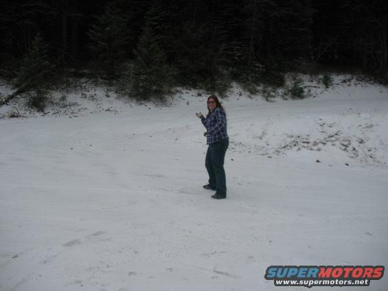 Woman stands in a snow-covered area with trees in the background. Watermark reads SUPERMOTORS