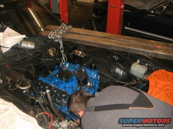 Mechanic working on a blue engine being lowered into a vehicle engine bay with chains and wooden beams