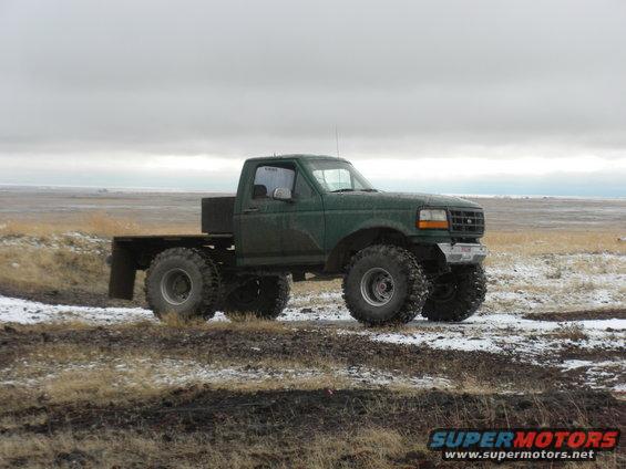 Green Ford pickup truck modified with large off-road tires and a flatbed, parked in a muddy, snowy field