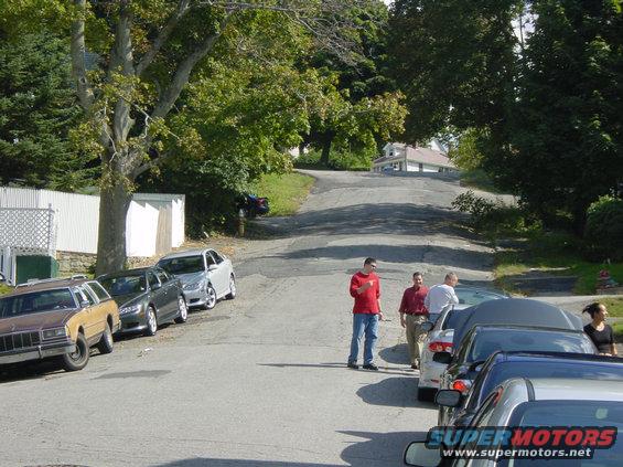 Brown station wagon, silver sedan, and dark sedan parked on a residential street with people standing nearby