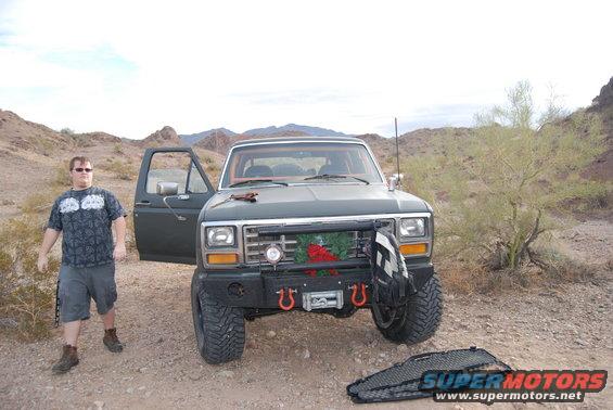 1980s Ford Bronco with open driver's door, man standing beside it, desert scenery. Aftermarket bumper with winch and tow hooks