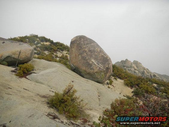 Large boulder resting on a rocky slope with sparse vegetation under a hazy sky