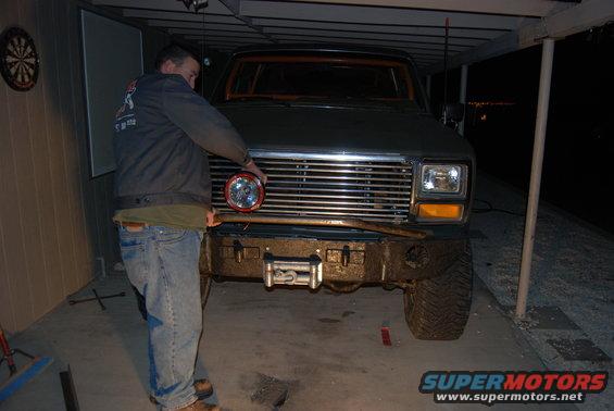 Dark green Ford Bronco front view with aftermarket bumper, winch, and a man installing a round headlight