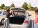 2003 Mazda 6 - A woman works on the trunk latch of a silver sedan while a man watches, with another car parked nearby.