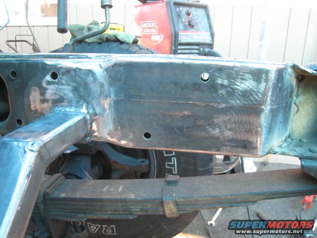 Truck frame with leaf springs and suspension components, showing welding and metal work. A Lincoln Electric welder is in the background