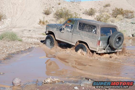 Dark green Ford Bronco splashing through mud, spare tire on rear