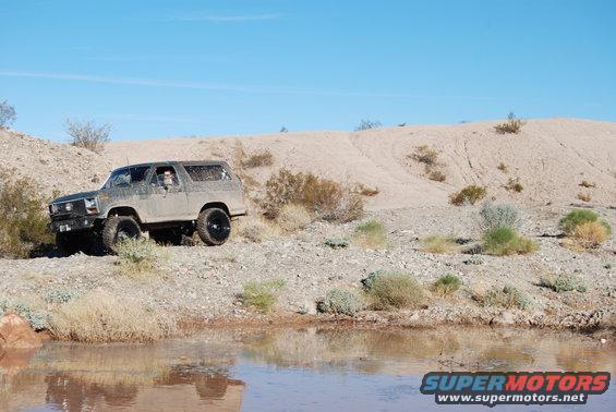 Ford Bronco with aftermarket wheels driving on a dirt path near a muddy puddle