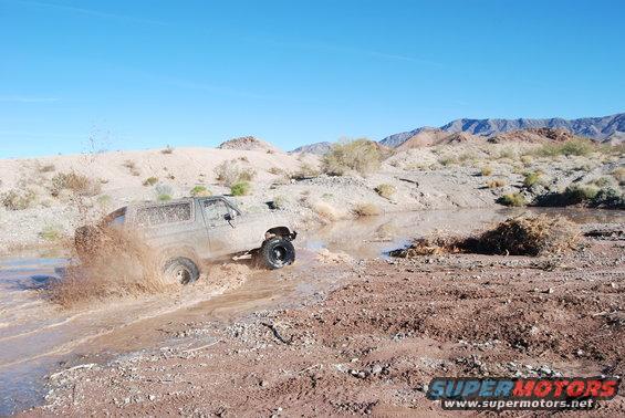 Ford Bronco kicking up mud while driving through a shallow stream in a desert landscape