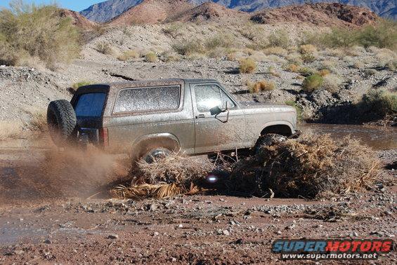 Silver Ford Bronco driving through mud and brush, spare tire on rear door, desert mountains in background