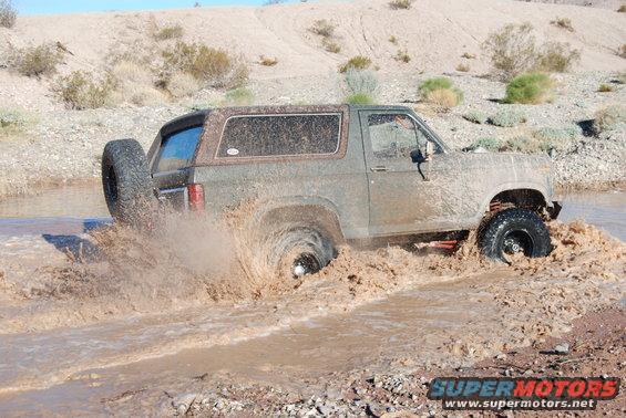 Ford Bronco driving through muddy water, kicking up spray. The vehicle is covered in mud