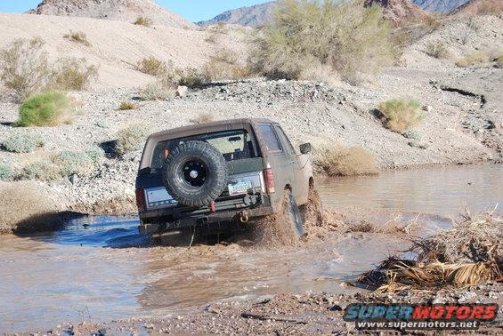 Brown Ford Bronco driving through muddy water, spare tire on rear door