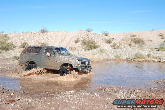 Muddy 1980s Ford Bronco splashing through a shallow water puddle in a desert landscape