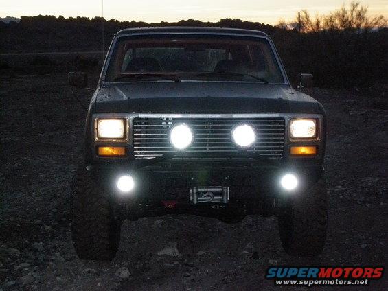Front view of a dark-colored Ford Bronco with two large round auxiliary lights mounted on the grille and two smaller ones below the bumper. A winch is visible between the lower lights
