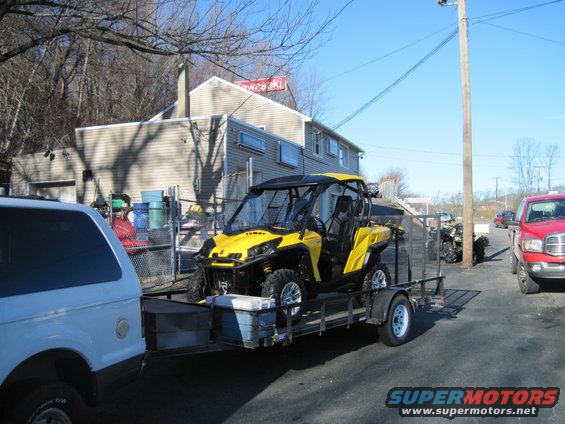 Yellow Can-Am side-by-side ATV loaded on a trailer, parked next to a white SUV and a red pickup truck
