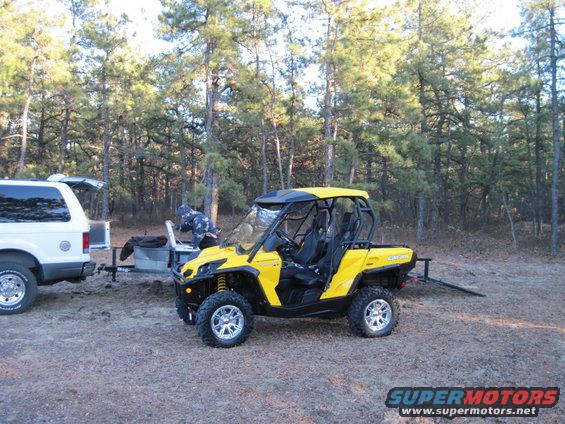 Yellow Can-Am ATV parked next to a white SUV with its trunk open. Trees in background