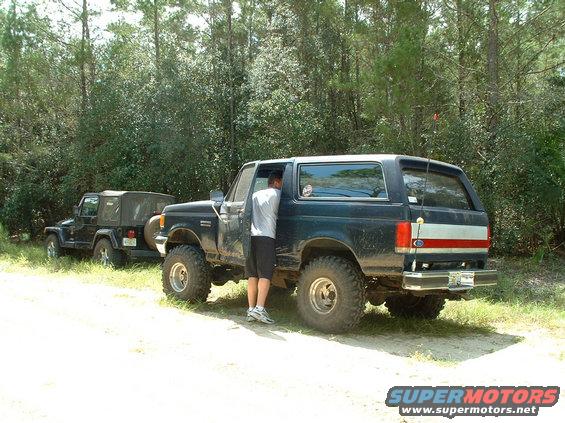Dark blue Ford Bronco with red and white stripe on rear, parked next to a black Jeep Wrangler. Person is leaning into Bronco
