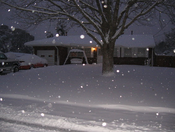 Snowy driveway with two cars covered in snow in front of a house