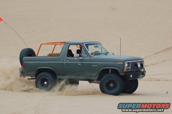 1982 Ford Bronco with orange roll cage driving through sand dunes, kicking up dust