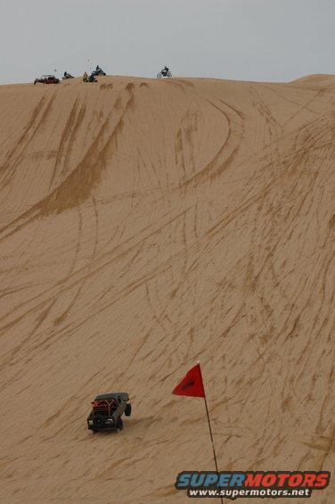 off-road buggy with roll cage driving down a sand dune, red flag nearby, other vehicles on top of dune