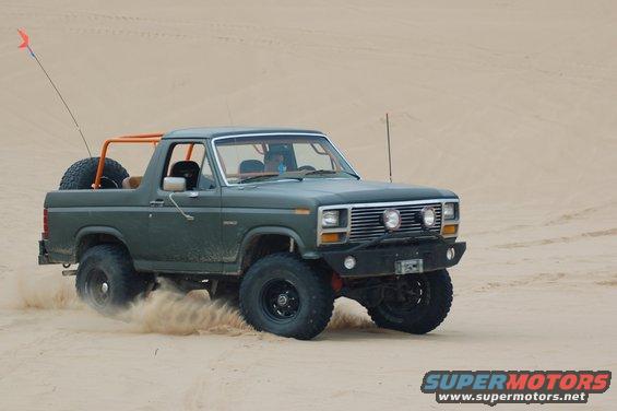 Green Ford Bronco with orange roll cage driving on sand, kicking up dust
