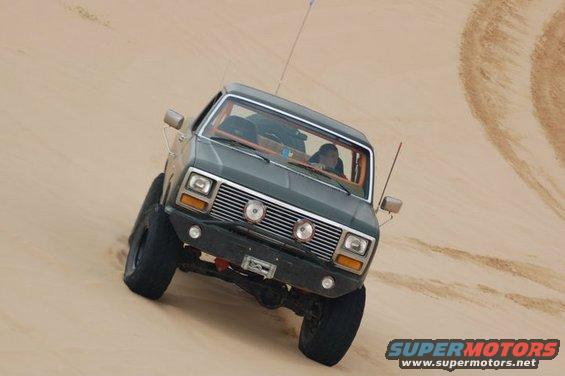 Dark green Ford Bronco with aftermarket bumper and winch driving up a sand dune. The vehicle has large tires and a CB antenna