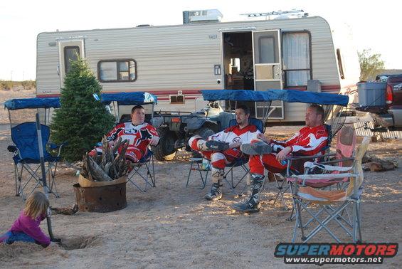 Three men in motocross gear sit in chairs around a fire pit near a camper and an ATV