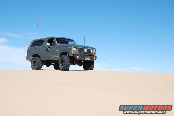 Green Ford Bronco with aftermarket bumper and large tires parked on a sand dune under a clear blue sky
