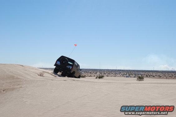 Bronco climbing sand dune with flag on antenna. Desert landscape with wind turbines in background