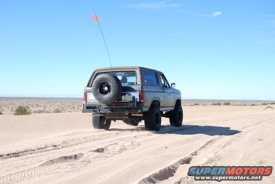 Gray Ford Bronco with spare tire on rear, driving on sand dunes under a clear blue sky