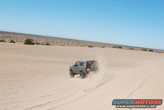 Green 1982 Ford Bronco driving on a sand dune, kicking up dust