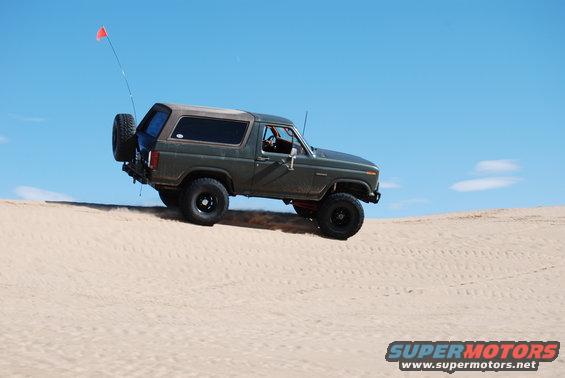 Green Ford Bronco with spare tire on back driving up a sand dune, red flag on antenna