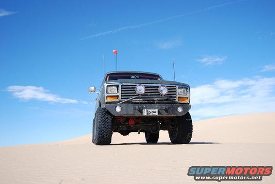 Front view of a lifted, dark green truck with off-road tires and a winch on the bumper, parked on a sand dune under a blue sky