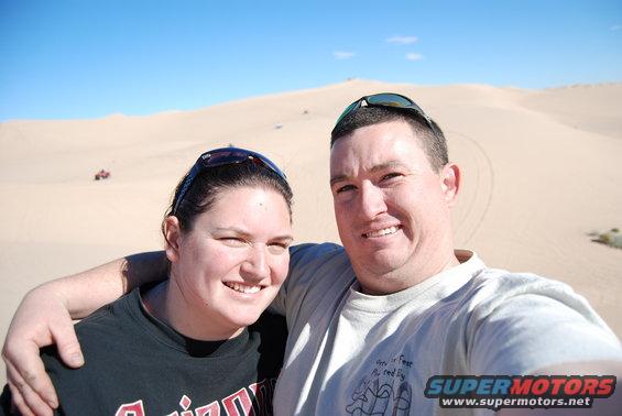 Two smiling people pose for a selfie in a desert with sand dunes under a bright blue sky. A small red ATV is visible in the distance