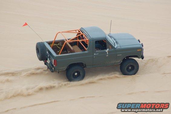 Olive green Ford Bronco with orange roll cage and spare tire on back driving in sand dunes