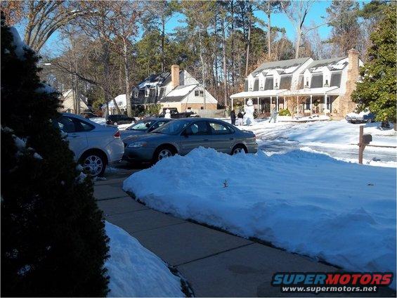Cars parked in a driveway covered in snow, with houses and trees in the background