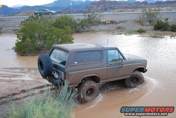 Muddy 1982 Ford Bronco driving through a shallow body of water with a spare tire on the back