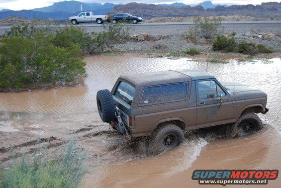 Mud-splattered brown Ford Bronco driving through a muddy puddle, with a spare tire mounted on the rear