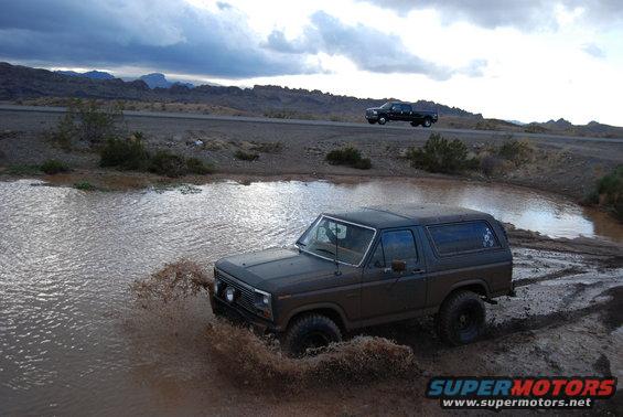 Dark 1980s Ford Bronco splashing through a muddy puddle off-road, with a black pickup truck driving on a road in the background