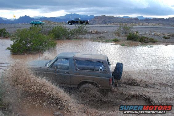 Ford Bronco splashing through a muddy puddle on a dirt road. Another truck and car are visible on the highway in the background