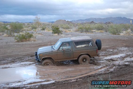 Mud-splattered olive green Ford Bronco stuck in a muddy puddle, rear-mounted spare tire visible