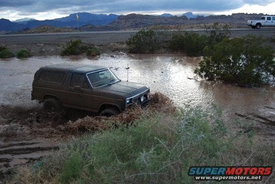 Brown Ford Bronco driving through muddy water with mountains in background