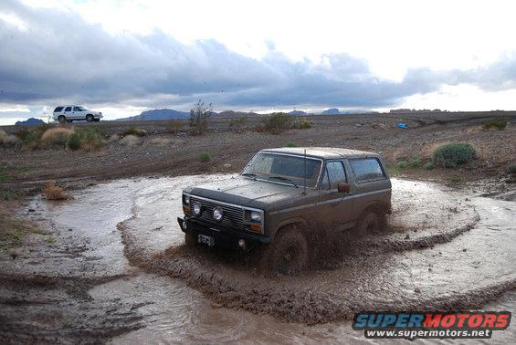 Brown Ford Bronco driving through mud, front-left 3/4 view. Another white SUV is visible in the background