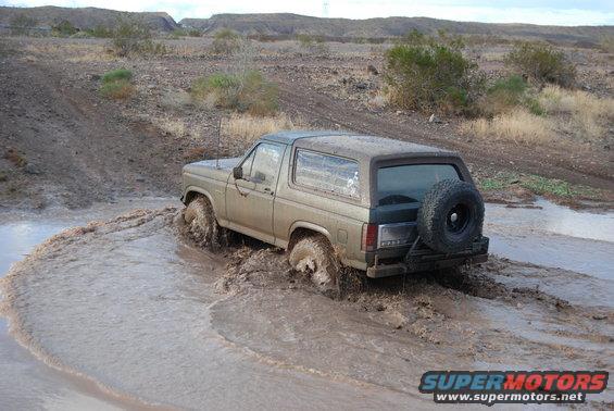 Tan Ford Bronco driving through mud, spare tire on rear door