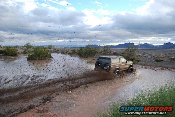 Tan 1982 Ford Bronco driving through mud, kicking up spray