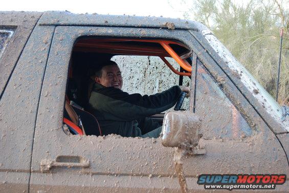 Mud-covered 1980s Ford Bronco interior with orange roll cage, driver smiling