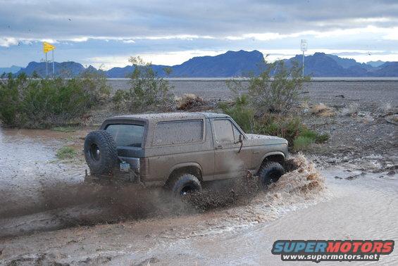 1982 Ford Bronco driving through muddy water, spare tire on back