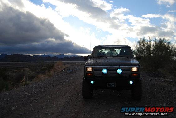 Front view of a dark-colored Ford Bronco with blue aftermarket lights on the grille and bumper, parked on a dirt road